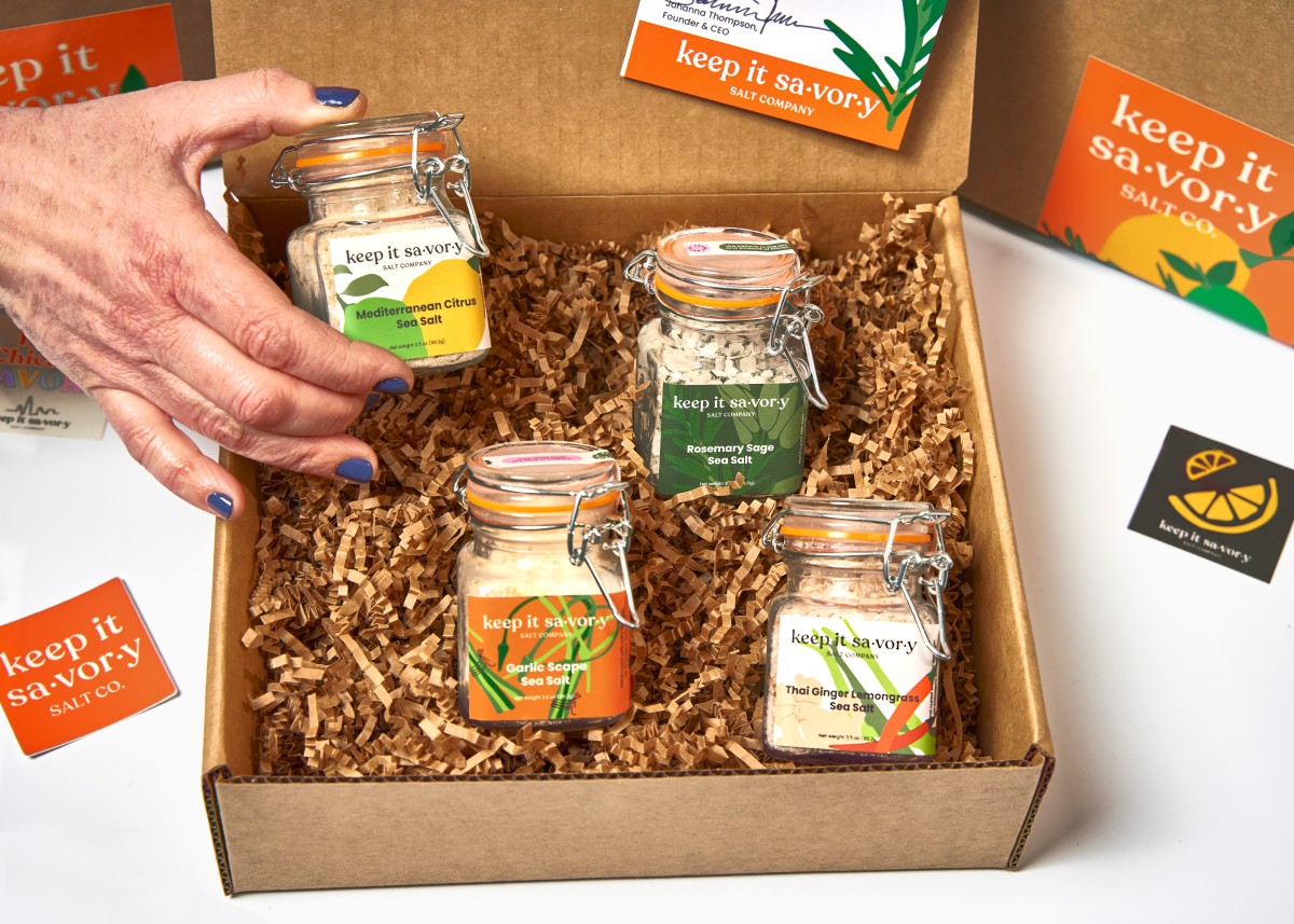 A gift box of Keep It Savory Salt Co. artisanal sea salts in glass jars with labels for Mediterranean Citrus, Rosemary Sage, Garlic Scape, and Thai Ginger Lemongrass, displayed on shredded paper packaging.
