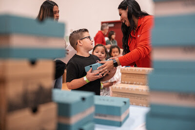 A smiling TOMS staff member hands a shoebox to a young student during a shoe distribution event at Monson-Sultana School, surrounded by stacked donation boxes.