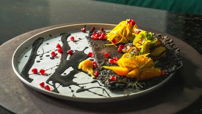 A beautifully plated dish featuring charcoal-crisped textures, bright mango and citrus elements, and scattered pomegranate seeds on an artistic black and white plate, representing Kimpton’s 2026 culinary trend forecast.
